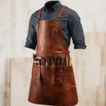 Brown Crazy Horse leather blacksmith apron worn over a dark shirt, featuring adjustable cross-back straps, chest pocket, multiple tool pockets, and durable handcrafted design for welding, woodworking, and metalworking tasks